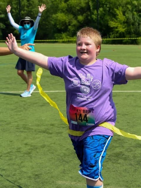 tri my best runner raises arms as he breaks the yellow finishline